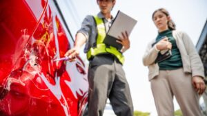 Insurance adjuster in yellow vest inspecting damaged red car with clipboard while woman stands nearby, assessing fault determination for accident claim.