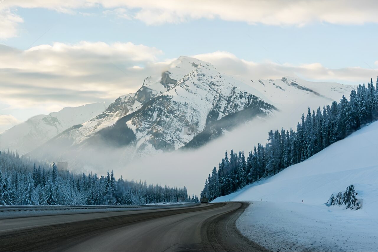 A scenic winter landscape featuring a winding road, snow-covered trees, and a misty mountain range.