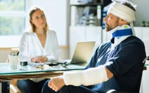 A man with a bandaged head, neck brace, and arm cast sits in an office, discussing with a woman at a desk with a laptop, emphasizing injury consultation.