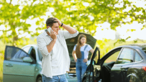 Man on phone looking stressed near a car crash, with woman by open car door, surrounded by green trees.