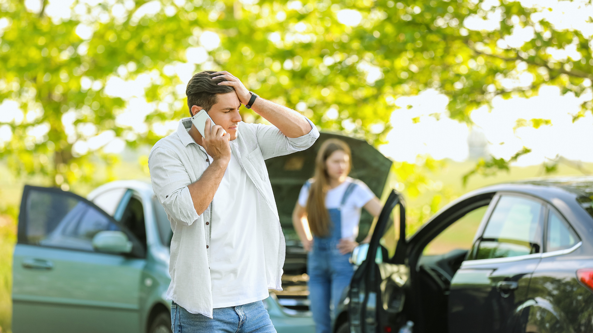 Man on phone looking stressed near a car crash, with woman by open car door, surrounded by green trees.