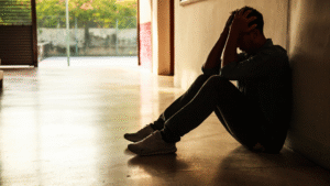 Silhouette of a person sitting against a wall with hands on head, showing distress in a hallway with light streaming in from an open door and greenery outside.