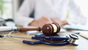 A wooden gavel and blue stethoscope on a desk, symbolizing a medical legal case, with a blurred person in a white coat in the background.