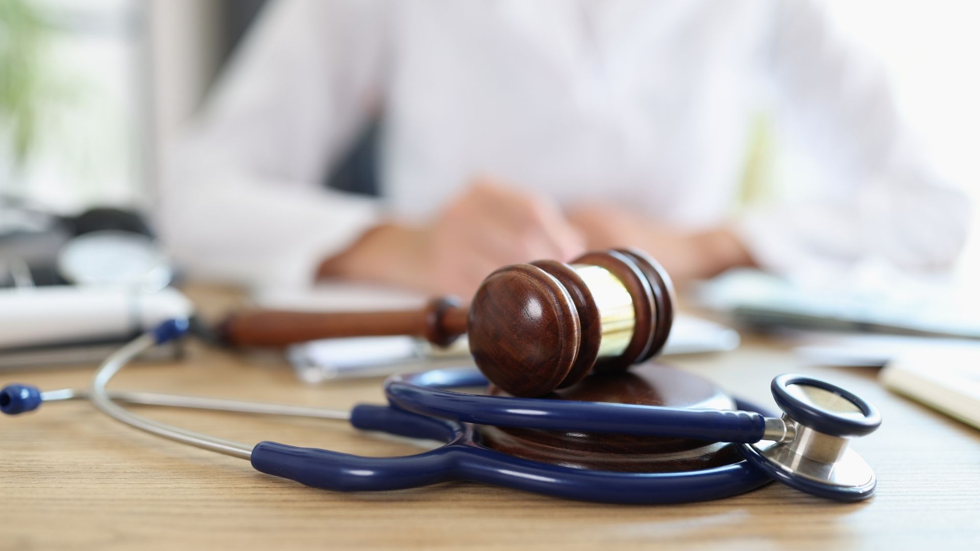 A wooden gavel and blue stethoscope on a desk, symbolizing a medical legal case, with a blurred person in a white coat in the background.