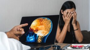 A woman with hands on face, distressed, next to a laptop showing a brain scan, with medical tools and pills on the table, highlighting brain injury concerns.