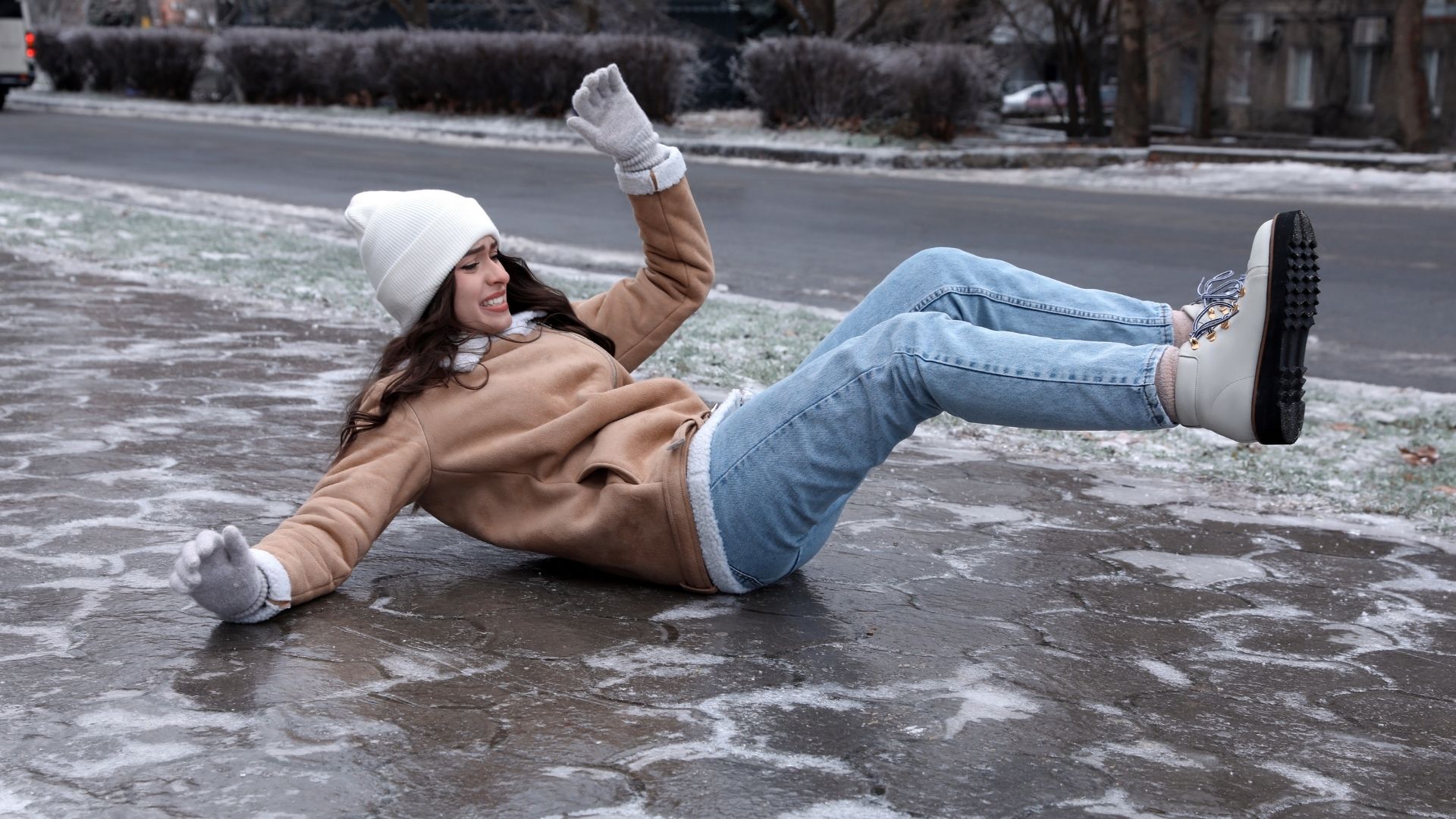 A person slipping on icy pavement, wearing a white beanie and gloves, highlighting the dangers of winter falls on slippery surfaces.