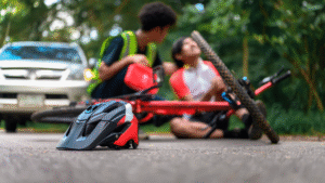 Cyclist injured on road with fallen bike and helmet, assisted by another person near a car, surrounded by greenery.