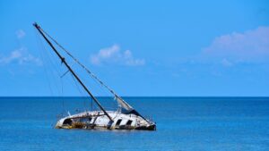 A partially sunken sailboat with a broken mast in calm blue ocean water under a clear blue sky, illustrating a boat accident settlement.