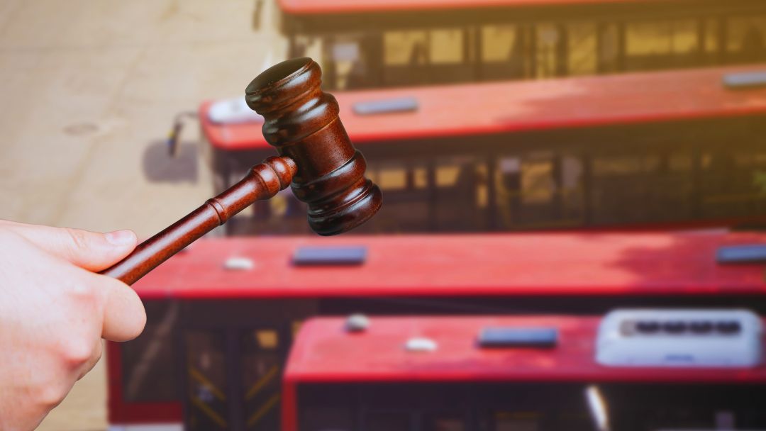 A hand holds a wooden gavel up against a blurred background of several parked red buses, symbolizing a bus accident settlement or legal decision.