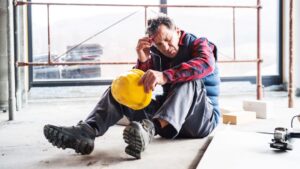 Distressed construction worker sitting on the floor of a site, holding his head and yellow hard hat, representing an injury claim.