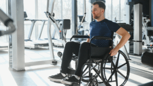 Man in a wheelchair smiling and looking forward while exercising in a brightly lit gym with weights and cardio machines in the background.
