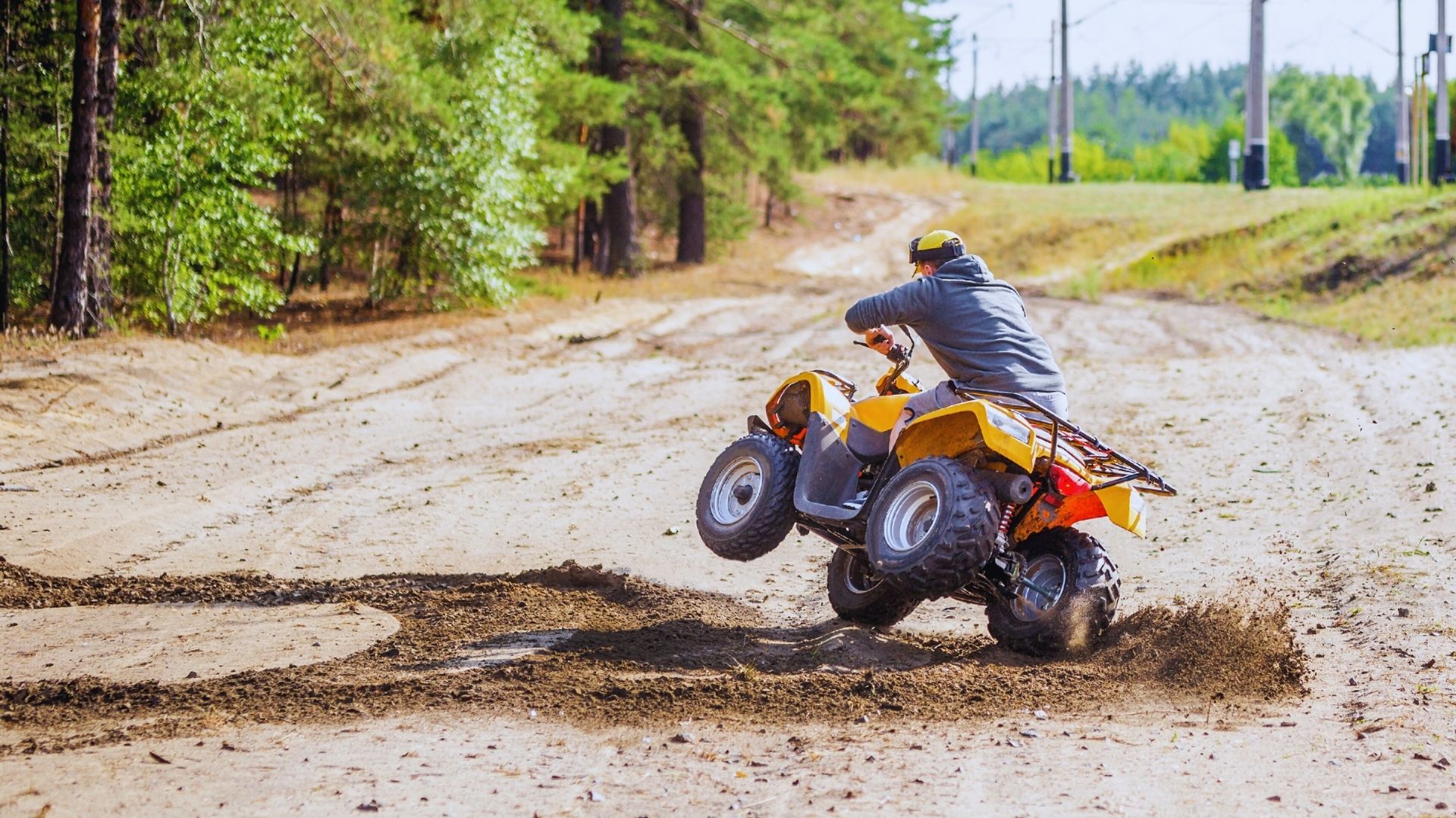 ATV rider in helmet and gray jacket on yellow quad bike navigating sharp turn on dirt trail in forested area, rear wheels airborne, spraying soil.