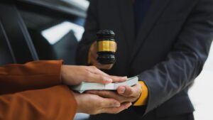 Hands exchanging cash with gavel near vehicle, representing insurance adjuster tactics car accident claims victims should understand after first contact.